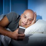 Bald Man Lying in Bed Looking at his Smart Phone Late at Night